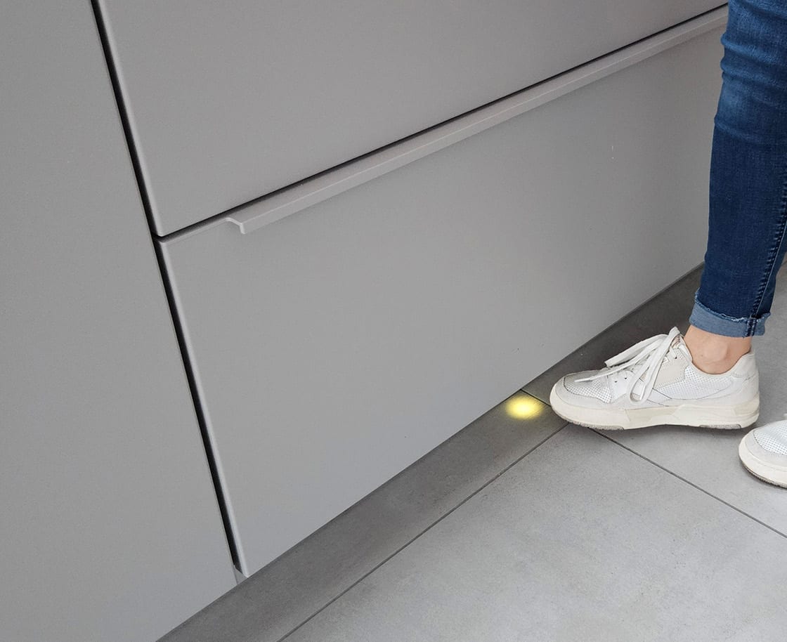 Un pied approche du socle d’un meuble bas gris ; un capteur logé dans la plinthe s’allume