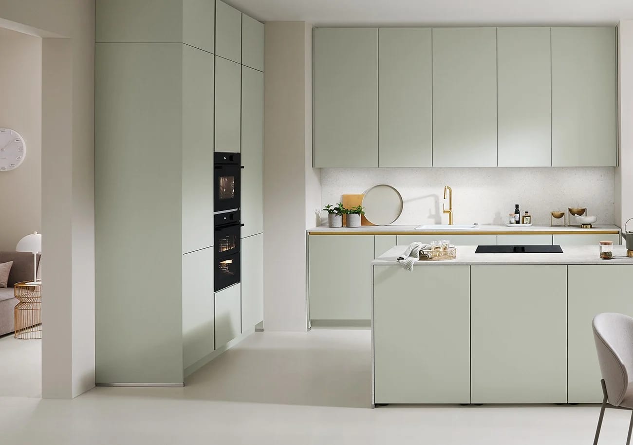 Cuisine claire aux façades vert d’eau mates : entre le plan de travail et les meubles hauts, une crédence pleine hauteur d’aspect terrazzo clair, éclairée par une ligne lumineuse