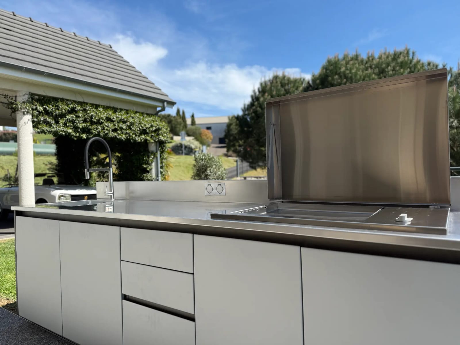 Plan de travail et cuve en inox d’une cuisine d’extérieur installée sur une terrasse, couvercle de plancha relevé