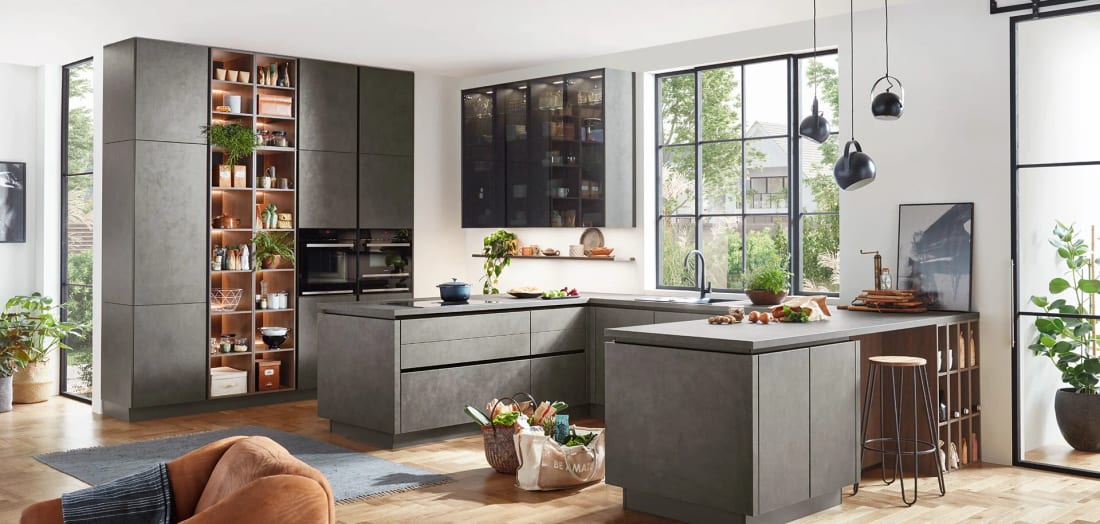 Cuisine en U en décor béton gris montée jusqu’au plafond, colonne de rangement ouverte et éclairée, meubles hauts vitrés