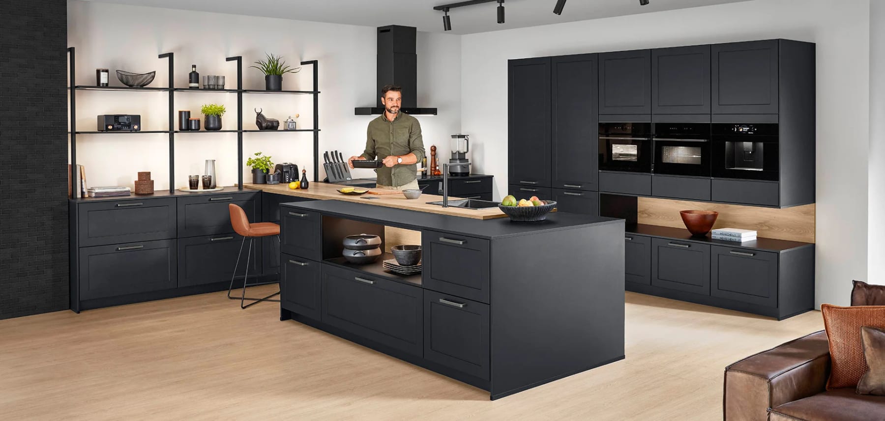 Cuisine aux façades à cadre noir mat : îlot central au plan de travail en bois, étagères sur montants métalliques et un homme qui prépare le repas